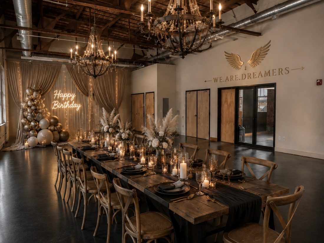 The venue set for a birthday party — long farmhouse table, candlelight, gold balloon arch and a 'Happy Birthday' neon sign