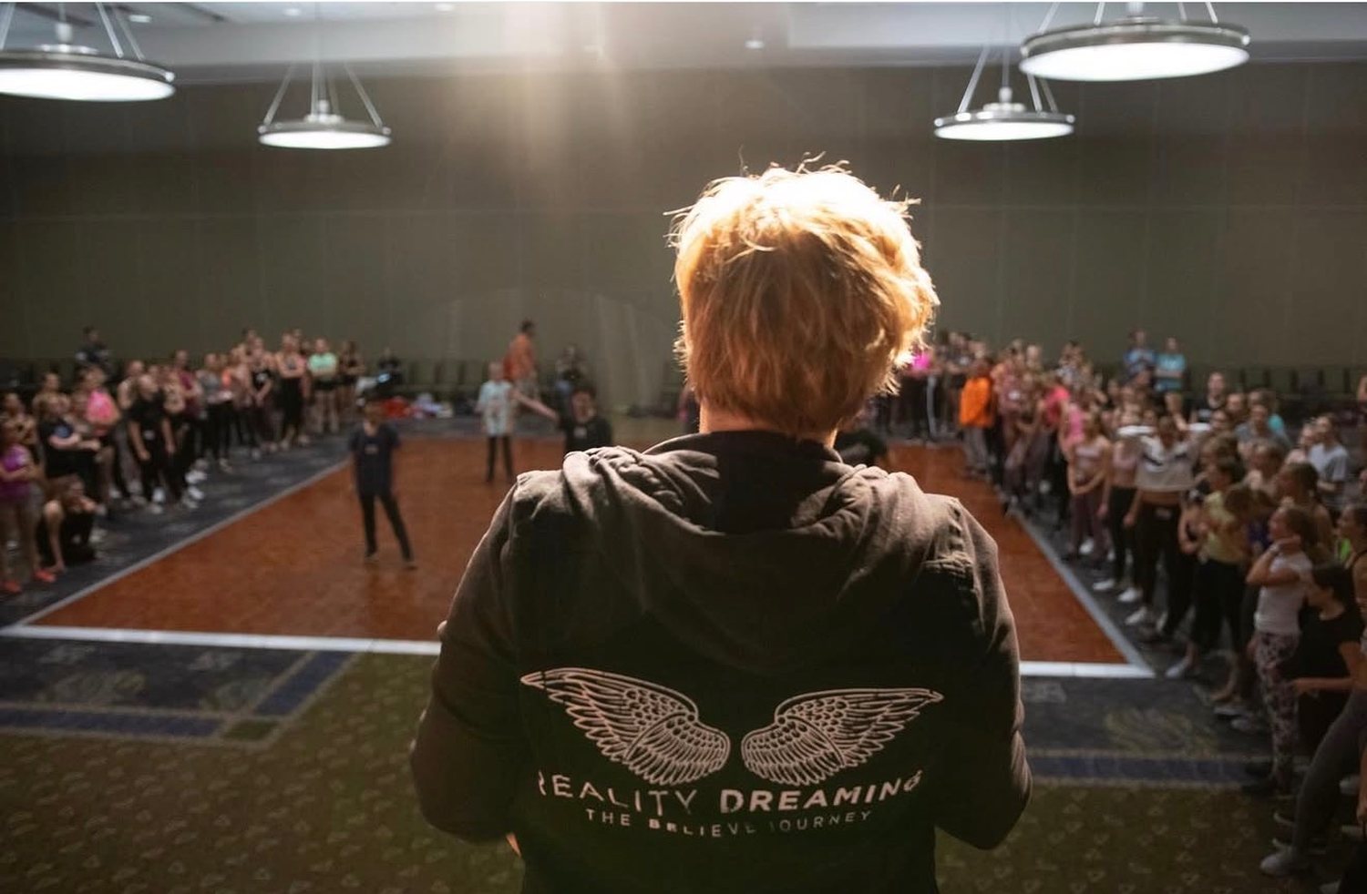 Rachelle addressing a full room of dancers, wearing a Reality Dreaming — The Believe Journey hoodie